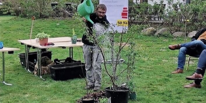 Der Veranstaltende gießt einen jungen Baum  mit einer grünen Gießkanne im Garten. Tische, Pflanzgefäße stehen im Hintergrund.