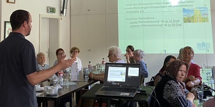 Der Blick geht vom Vortragenden in Richtung Bürger*innen und Präsentationsleinwand. Das Laptop steht aufgeklappt dazwischen. Auf den Tischen stehen Getränke und Gläser, Tassen.