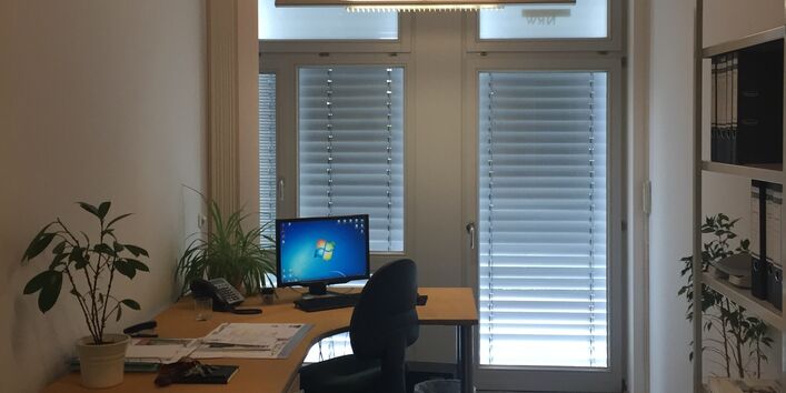 Ein Büro mit Schreibtisch und Stuhl mit zwei großen Fenstern und heruntergelassenen Sonnenrollos