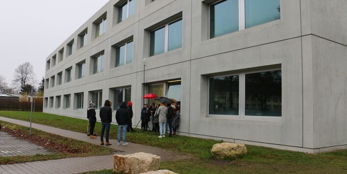 Vor einem grauen Gebäude stehen mehrere Personen in einer Gruppe um ein Messgerät.