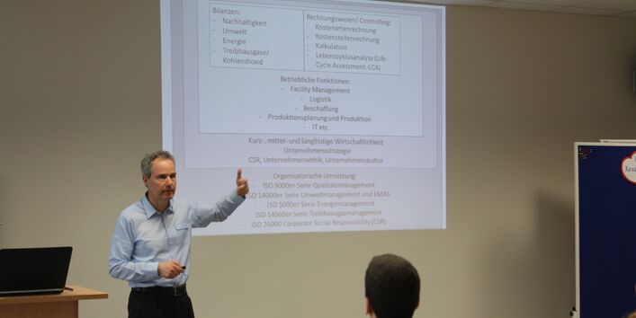 Vortragender steht gestikulierend vor Publikum. An seiner linken Seite stehen zwei Moderationswände, an seiner rechte Seite befindet sich ein Rednerpult. Auf dem Rednerpult liegt ein aufgeklapptes Laptop.