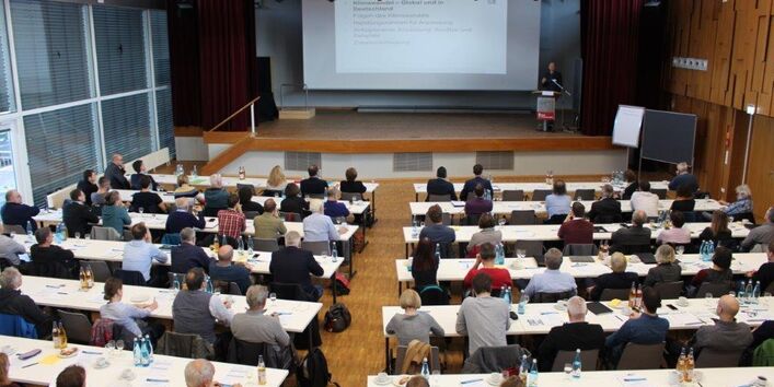 Von hinten oben wurde eine Halle fotografiert, in der an Tischen, die zu einer Bühne ausgerichtet ist, viele Menschen sitzen. Auf der Bühne hält ein Sprecher eine Präsentation.