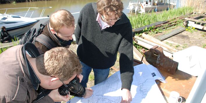 3 Männer stehen am Wasserlauf in der Sonne und schauen über eine auf dem Tisch ausgebreitete Karte. Ein Mann zeigt auf die Karte, wobei die anderen beiden Personen fotografieren und zusehen.