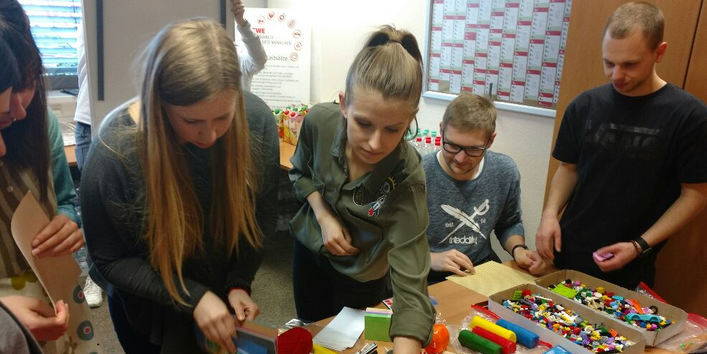 Personen schauen auf einen Tisch mit bunten Moderationsmaterialien und Stiften und greifen danach.