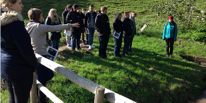 Personen stehen an einer mit Gras bewachsenen Böschung. Im Hintergrund grasen Schafen. Im Vordergrund zeigt eine Teilnehmerin mit gestreckten Arm in eine Richtung; eine andere Teilnehmerin steht hinter einem Holzzaun. Alle Personen schauen in die Richtung des gestreckten Armes.