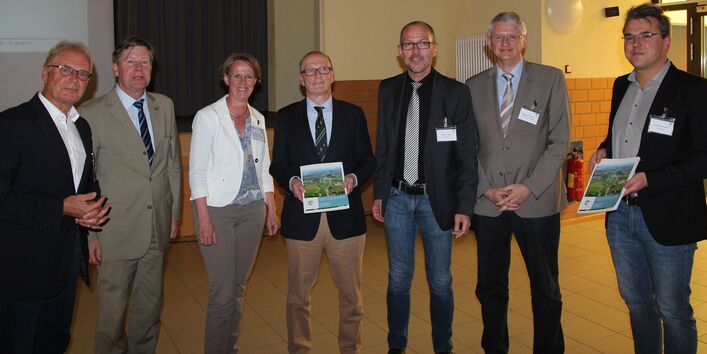 Gruppenbild von Personen, 7 Männern, 1 Frau. Zwei Männer halten einen Bericht präsentierend in der Hand.