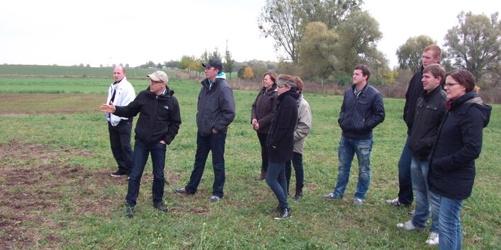 Personen stehen auf einem Rasen schauend in Richtung von Versuchsfeldern und hören dem Fachreferenten zu.  Im Hintergrund befinden sich wenig belaubte Bäume. Der Himmel ist grau.