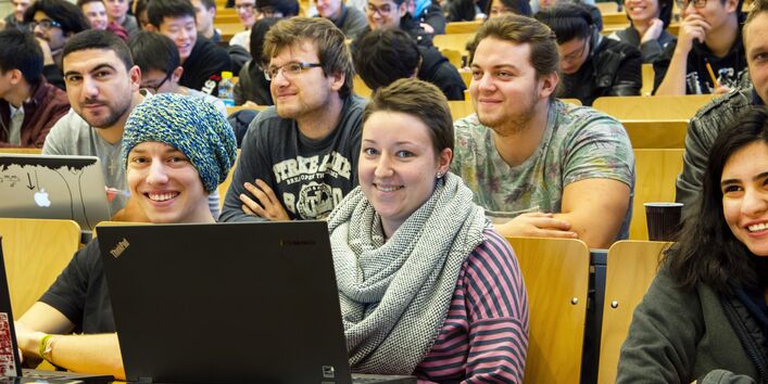 In einem Hörsaal sitzen Studierende, manche mit aufgeklapptem Laptop vor sich. Zwei Personen in der ersten Reihe schauen in die Kamera, andere schauen nach vorne und lachen.