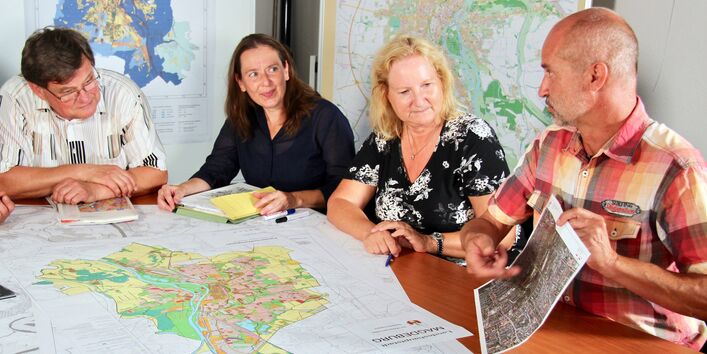 An einem Tisch, auf dem mehrere Karten liegen, sitzen mehrere Personen. Eine Person deutet auf eine kleineren Karte, die sie in der Hand hält.