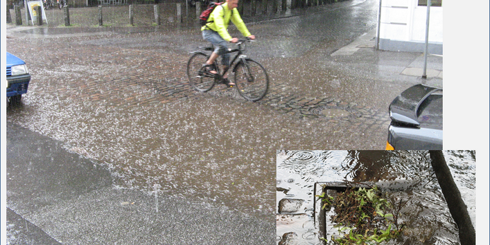 Ein Mann in einem hellgrünen Pullover und mit Rucksack auf dem Rücken fährt mit seinem Fahrrad auf einer Straße mit Pflastersteinen ungeschützt durch besonders starken Regen. Im Hintergrund sieht man einen Ausschnitt von einem Backsteingebäude mit Fahrrädern und Fahrradständer im Vordergrund, begrenzt durch Natursteinpoller zur Straße hin. Die Straßen werden durch Bäume und Autos gesäumt. Im Vordergrund rechts unten sieht man das Bild eines vom Regen mit Pflanzenresten zugeschwemmten Gulli.