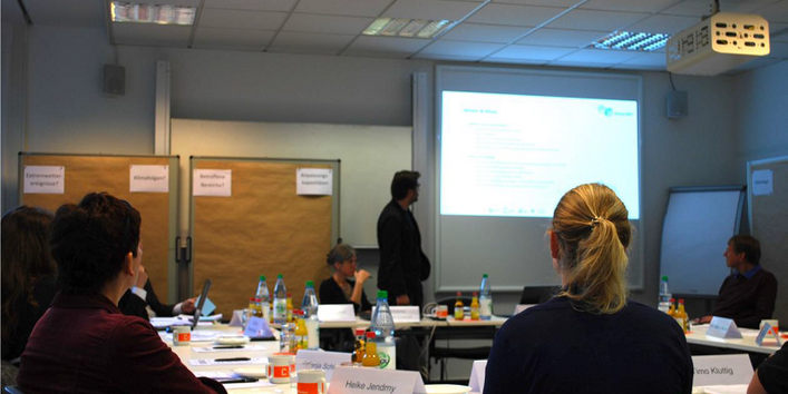 Teilnehmende sitzen an Tischen verteilt in U-Form und schauen auf die Präsentation an der Projektionsfläche an der Wand. Der Raum ist halb verdunkelt. Auf den Tischen liegen Arbeitspapiere und es stehen Getränkeflaschen und Tassen, Namensschilder darauf. Im Raum stehen rundum Moderationswände verteilt.