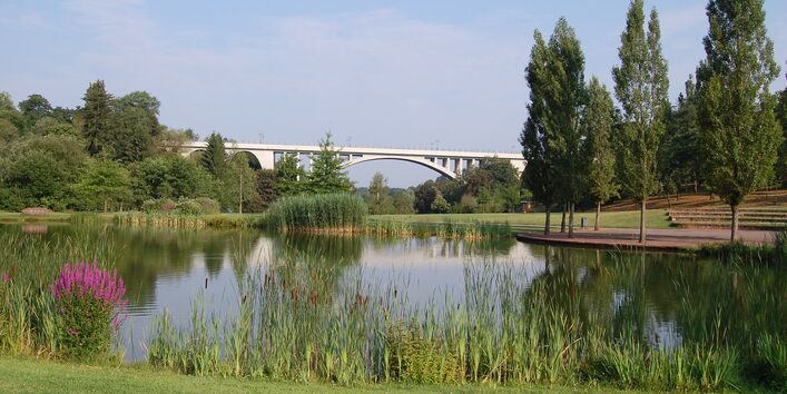Der Blick geht auf eine weisse Brücke im Hintergrund vor blauem Himmel. Im Vordergrund spiegelt sich das Bild im Wasser, welches von Bäumen gesäumt ist.