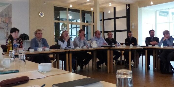 Seminarteilnehmende sitzen an Tischen in U-Form im Seminarraum. Ein großes Fenster seitlich im Hintergrund lässt den Raum lichtdurchflutend wirken.