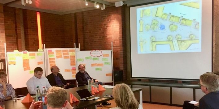 Personen sitzen am Tisch und tauschen Ideen zum Plangebiet aus, welches an eine Leinwand projiziert ist