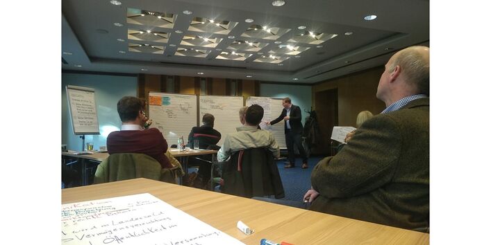 In einem Raum sitzen mehrere Personen zu Pinnwänden am Ende des Raumes gerichtet. Dort deutet eine Person auf eine Pinnwann und erklärt etwas.