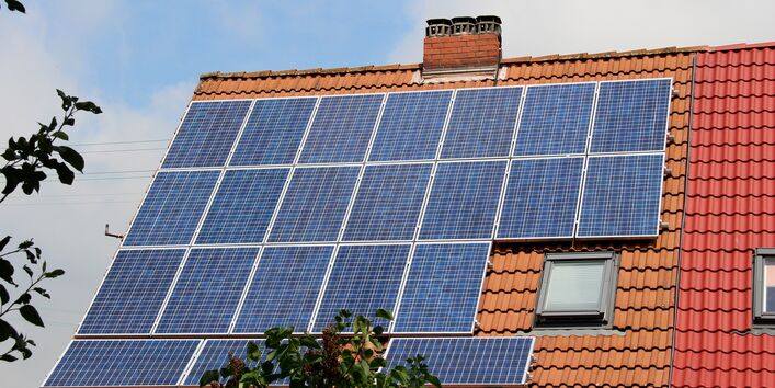 Der Blick geht auf ein rotes Ziegeldach, welches überwiegend mit Solarpaneelen bestückt ist. Im Hintergrund ist bewölkter Himmel zu sehen.
