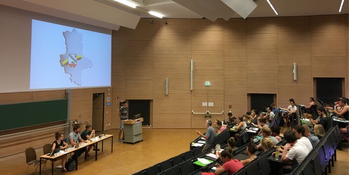 Blick in einen großen Hörsaal. Vorne sitzen drei Personen an einem Tisch, daneben eine Person an einem Rednerpult. Ihnen gegenüber sitzen weitere Personen in den Hörsaalrängen.