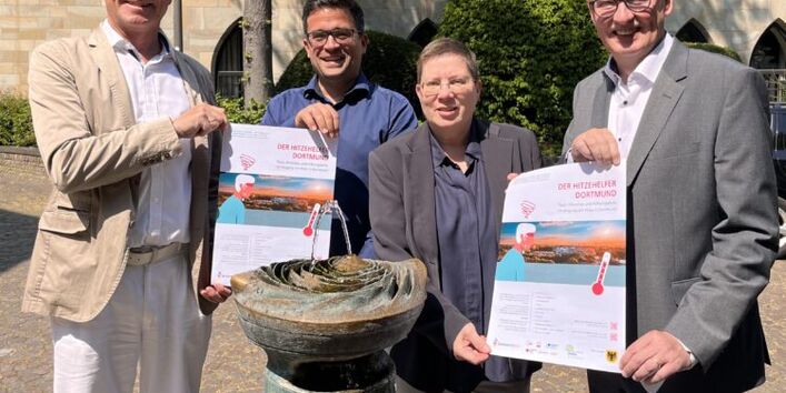 Das Bild zeigt vier Personen mit der Hitzehelfer-Broschüre vor einem Trinkbrunnen. 