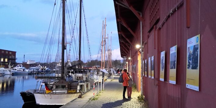 Passant*innen betrachten barrierefrei zugängliche Plakate der Ausstellung ÜberFlut an einem Schuppen an der Untertrave, Lübeck.