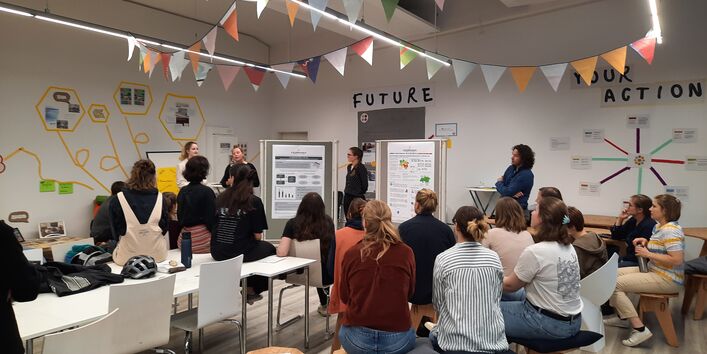 Posterpräsentation im mit Wimpeln ausgestatteten Raum. Der Blick geht über die sitzenden Teilnehmenden hinweg.