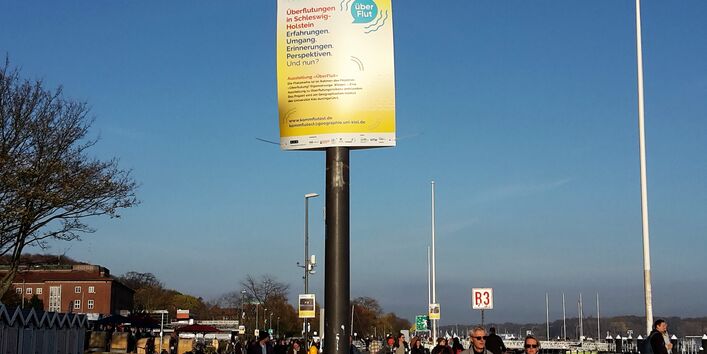 Plakate der Open-Air-Ausstellung ÜberFlut hängen barrierefrei an Laternenpfählen an der Kiellinie in Kiel.