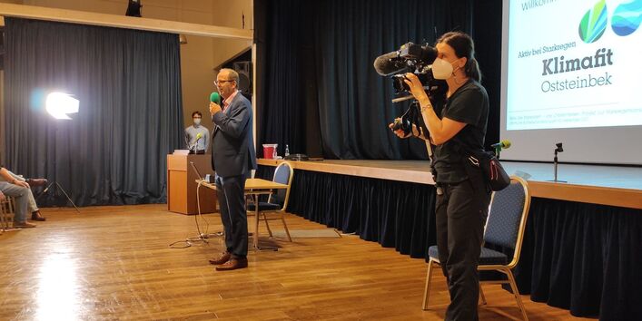 Ein Vortragender und eine Kamerafrau stehen vor der Bühne vor Publikum. Im Hintergrund ist das Willkommensbild zu sehen.