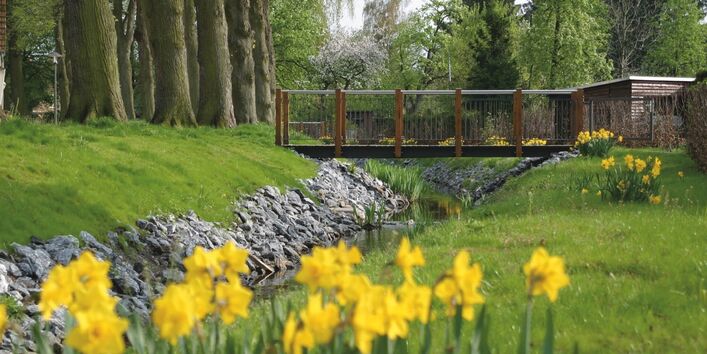 Ein Bach fließt unter eine Holzbrücke entlang von Bäumen und blühender Wiese.