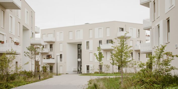 Der Blick geht auf die Außenanlagen im Quartier. Weiße quadratischen Wohngebäudekomplexe stehen in einer begrünten Wohnanlage.