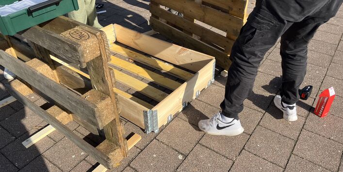 Zu sehen sind zwei Personen, die aus Paletten ein Hochbeet bauen.