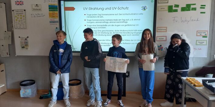 5 Kinder stehen vor einer digitalen Tafel in einem Klassenraum und stellen Ergebnisse ihrer Untersuchungen vor.