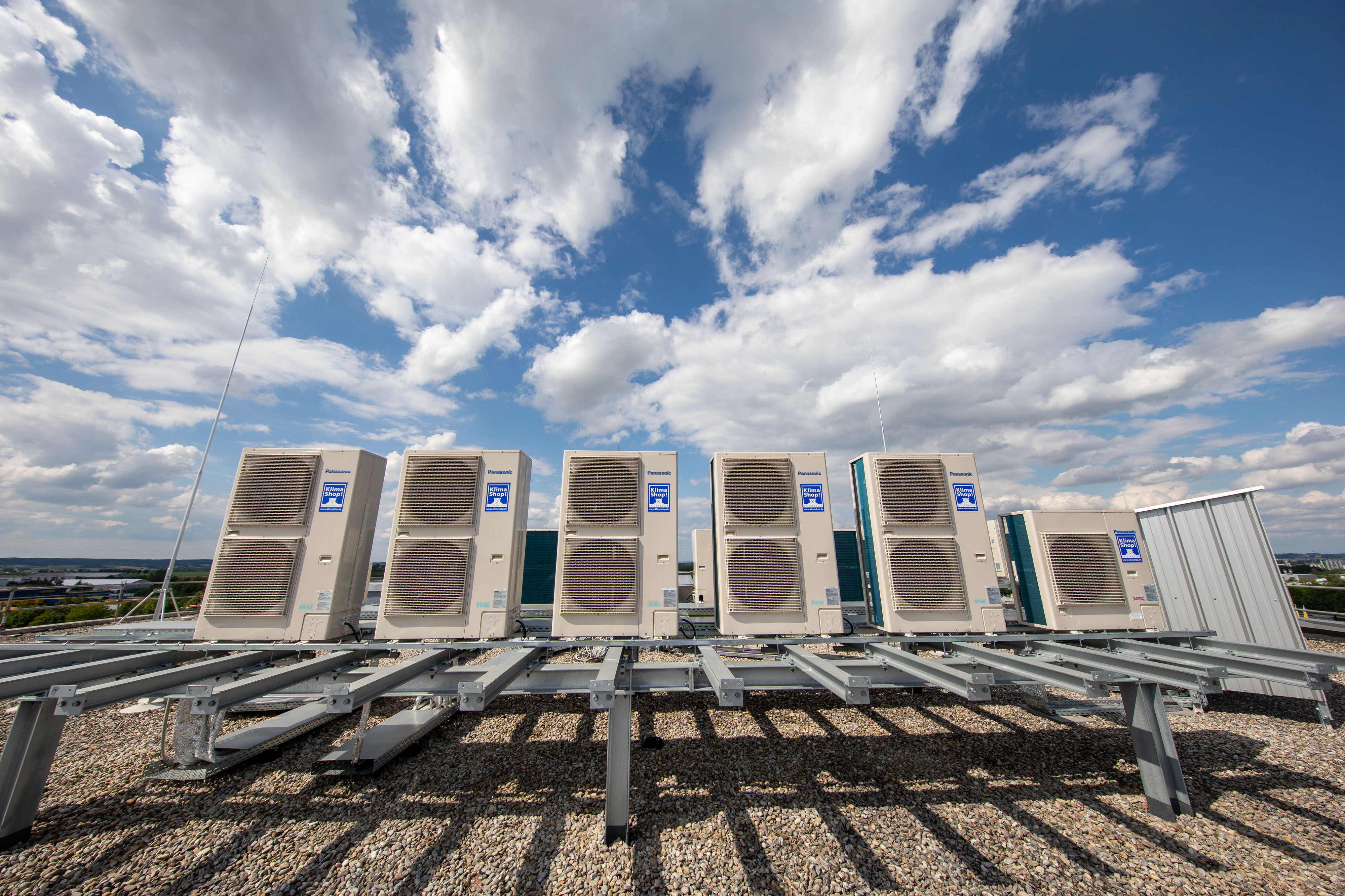 Die Komplettansicht der Außengeräte der Luft-Luft-Wärmepumpen auf dem Kiesdach vor sonnig-wolkigem Himmel