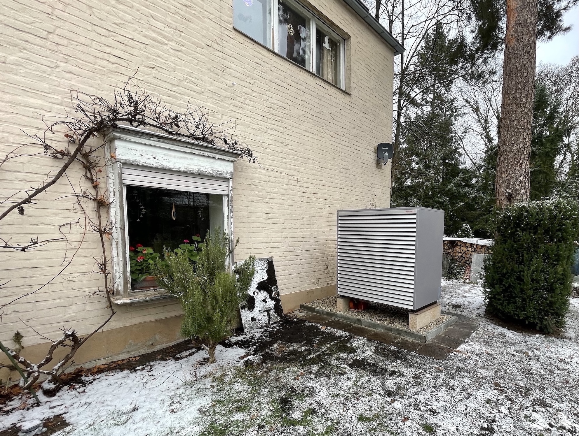 Ein Außengerät einer Wärmepumpe im leicht verschneiten Garten, quer zur beigen, geziegelten Hauswand aufgestellt.