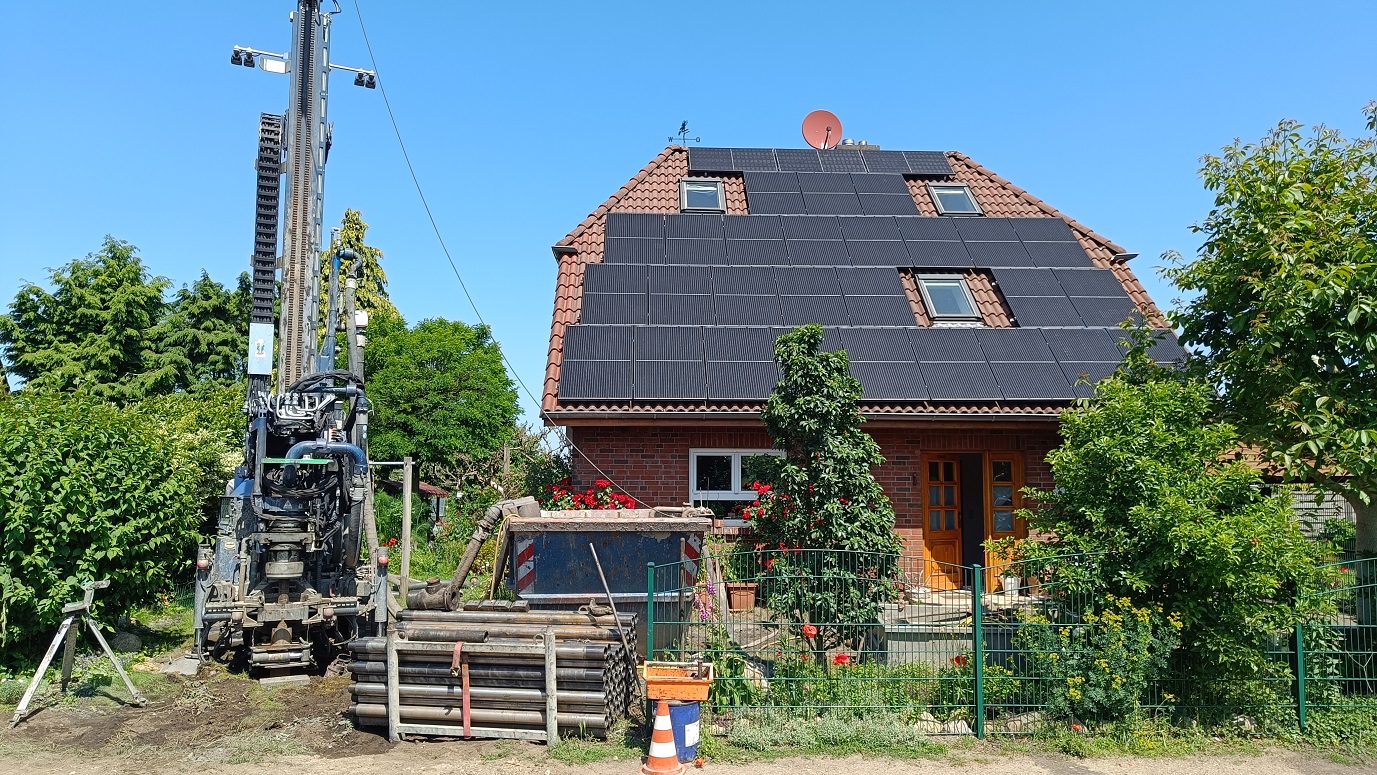 Ein geklinkertes Haus steht quer zur Straße, das Dach ist mit Photovoltaik belegt. Vor dem Haus im Vorgarten steht ein hohes Bohrgerät, daneben Stangen auf einer Palette und ein Container.