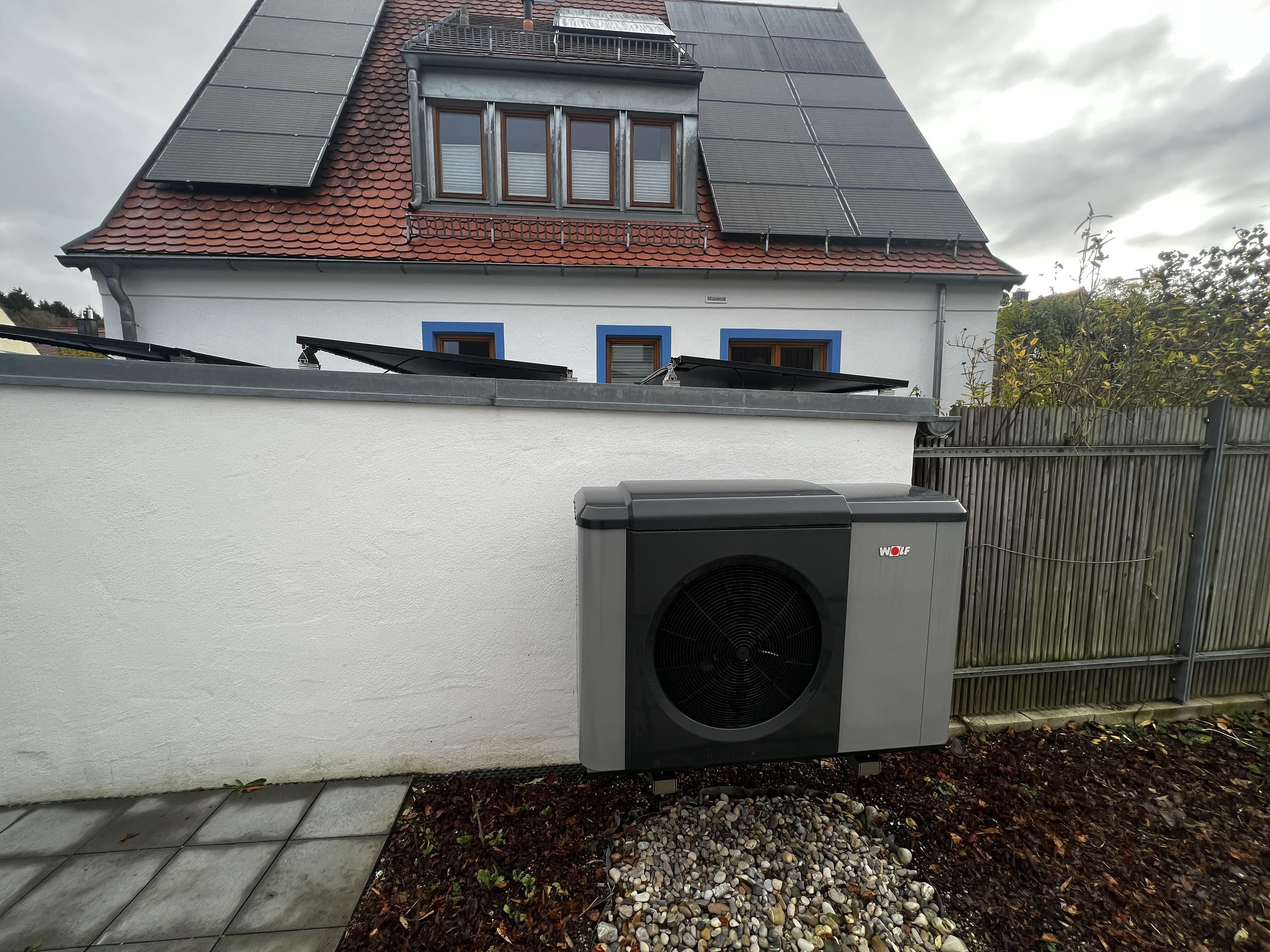 Eine Wärmepumpe an einer Grundstücksmauer, im Hintergrund das Einfamilienhaus.