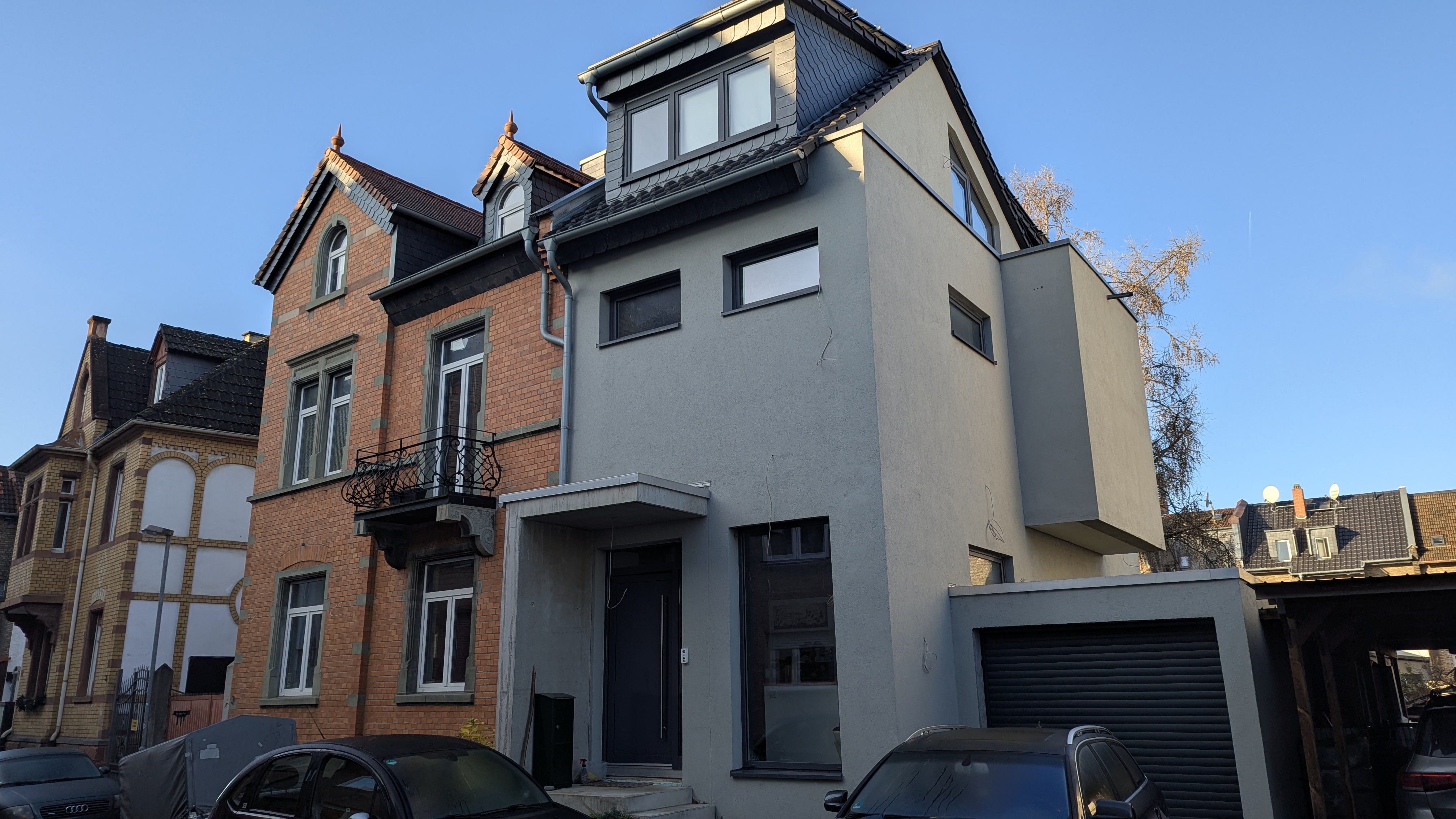 Die Fassade eines zweigeschossigen, rot geklinkerten Altbaus mit einem modernen, grau verputzten Anbau.